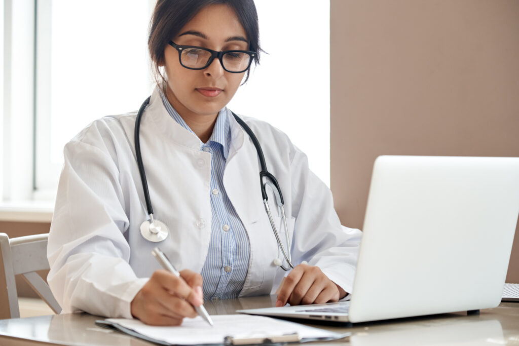 Indian ethnic female doctor physician gp wearing glasses, white coat stethoscope writing filling medical form watching online medical webinar seminar training working sitting with laptop at workplace.