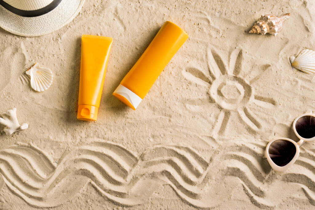 Summer travel beach flat lay composition. Wicker hat, sunglasses, sunscreen lotion, sunblock cream and seashells on beach sand background. Summer vacations and spf uv-protecting skin care concept.