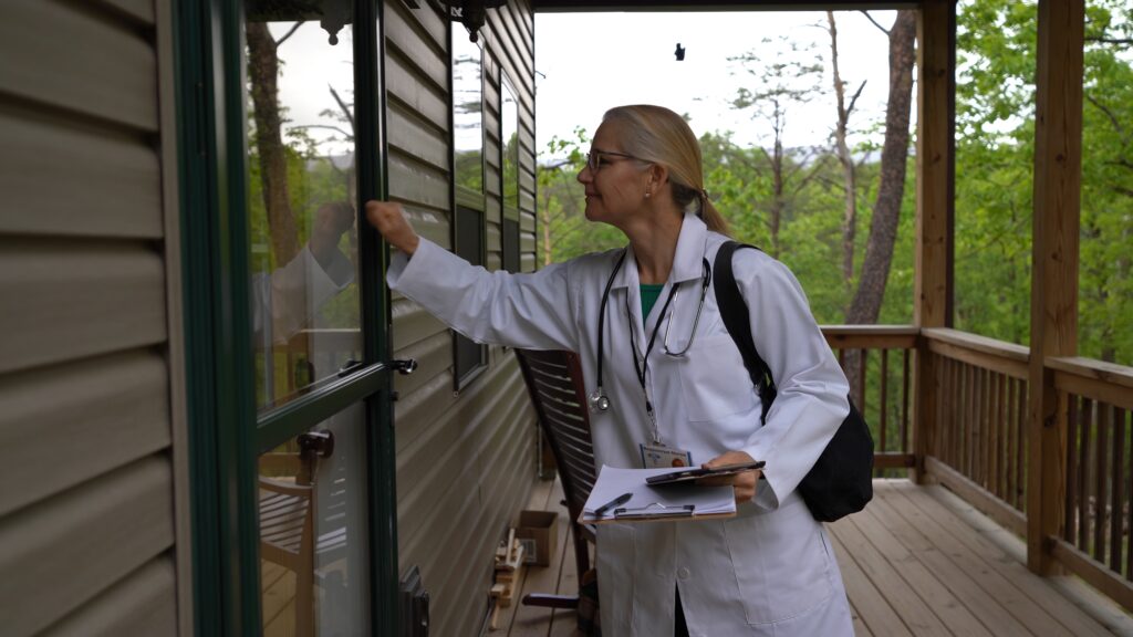 Medium wide shot of side view of nurse or doctor arriving for home visit in rural area, knocking and smiling at door.